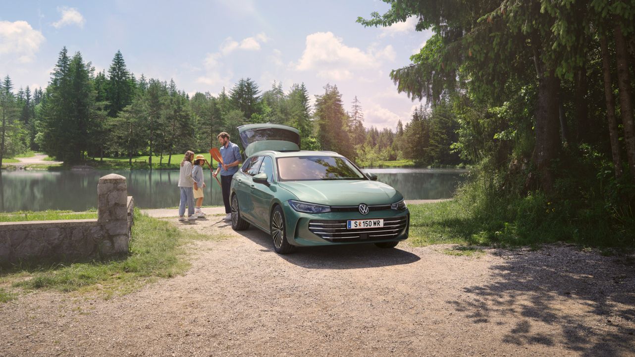 Der neue VW Passat Elegance in Maripositgrün Metallic parkt an einem Badesee mit geöffnetem Kofferraum. Ein Mann verteilt Freitzeitspielzeug an zwei Kinder.