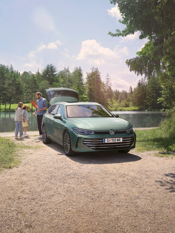 Der neue VW Passat Elegance in Maripositgrün Metallic parkt an einem Badesee mit geöffnetem Kofferraum. Ein Mann verteilt Freitzeitspielzeug an zwei Kinder.
