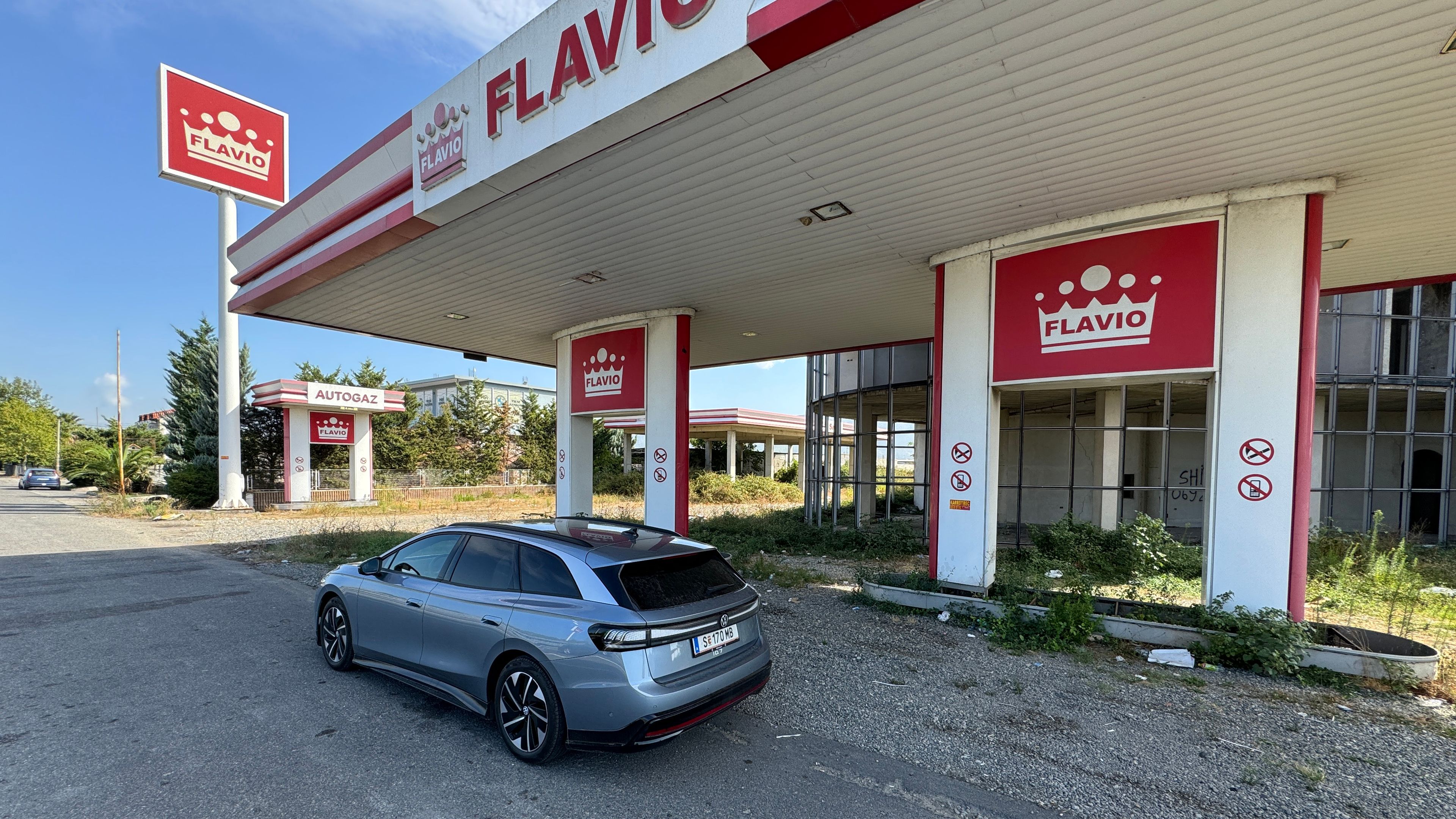 Ein grauer Kombiwagen parkt vor einer verlassenen Tankstelle mit roter Beschilderung, die den Namen "Flavio" trägt. Das Gebäude zeigt Anzeichen von Vernachlässigung.