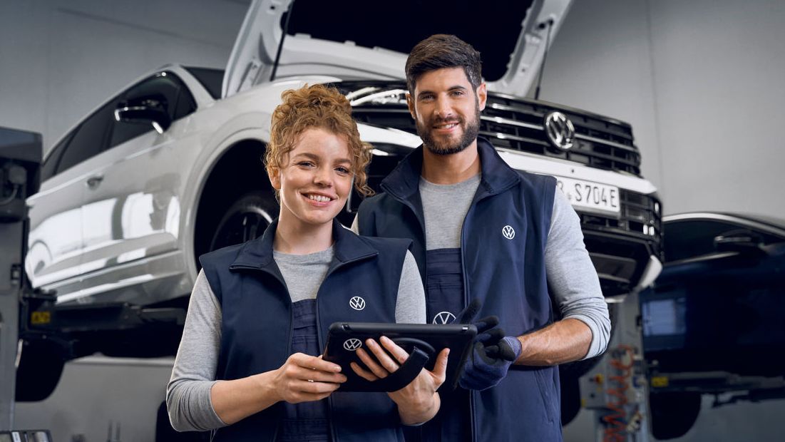 Zwei Kfz-Mechaniker:innen mit VW-Logos auf ihrer Arbeitskleidung stehen in einer Werkstatt vor einer Hebebühne auf der ein weißer Volkswagen Touareg mit geöffneter Motorhaube zu sehen ist. 