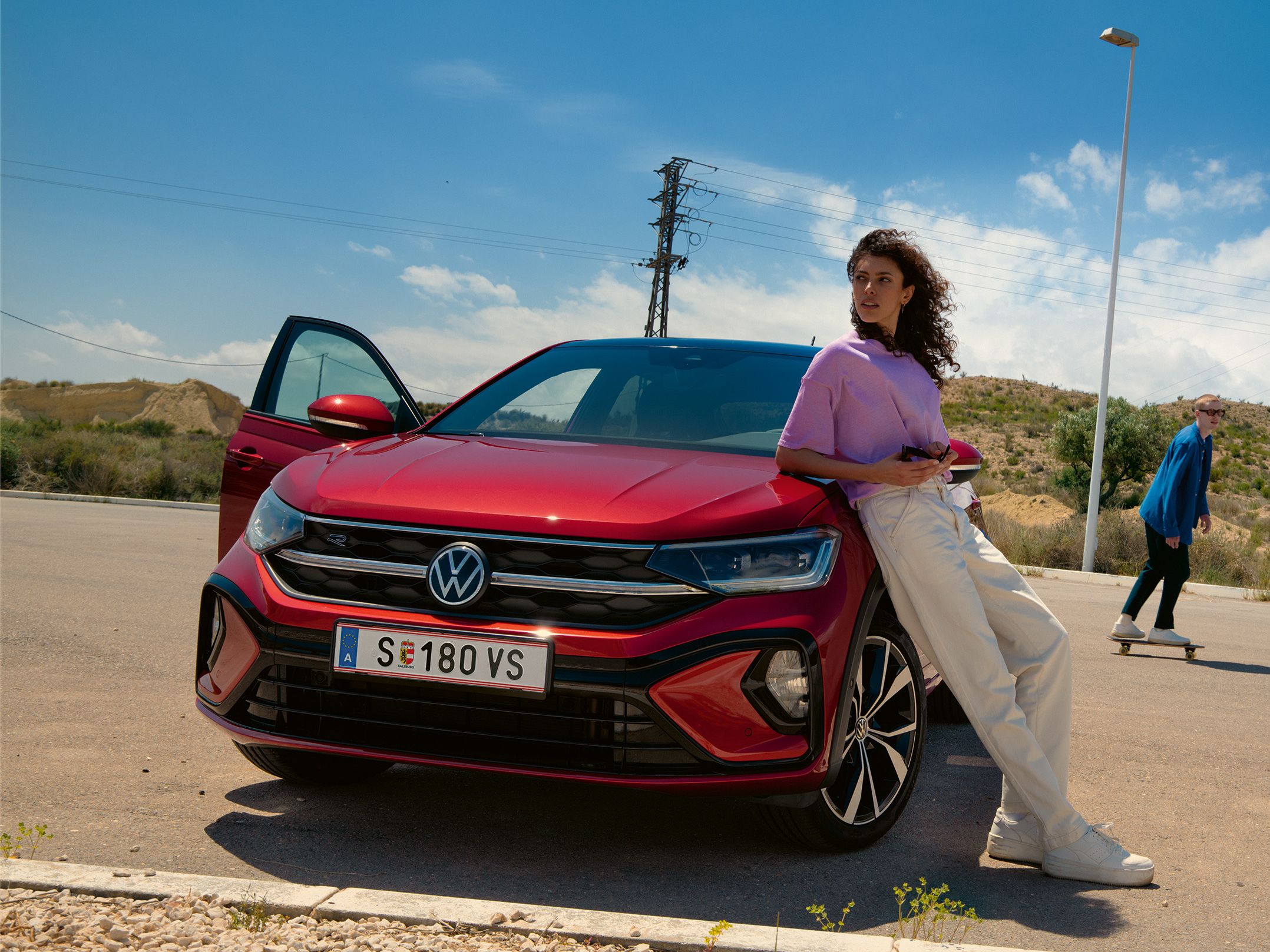 VW Taigo R-Line in Rot, Frontansicht auf Parkplatz. Frau lehnt an Motorhaube, Mann fährt Skateboard