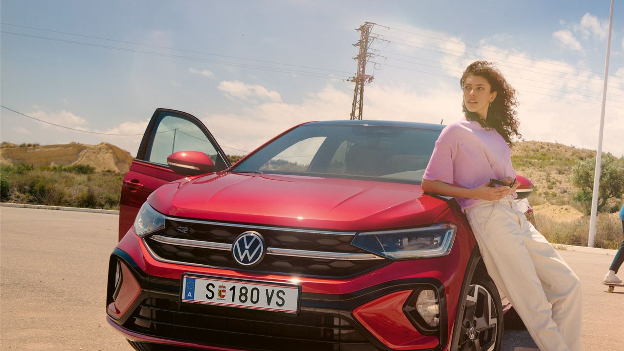 Frau lehnt an der Motorhaube eines VW Taigo auf einem Parkplatz in der Wüste. Mann fährt auf Skateboard dahinter entlang.