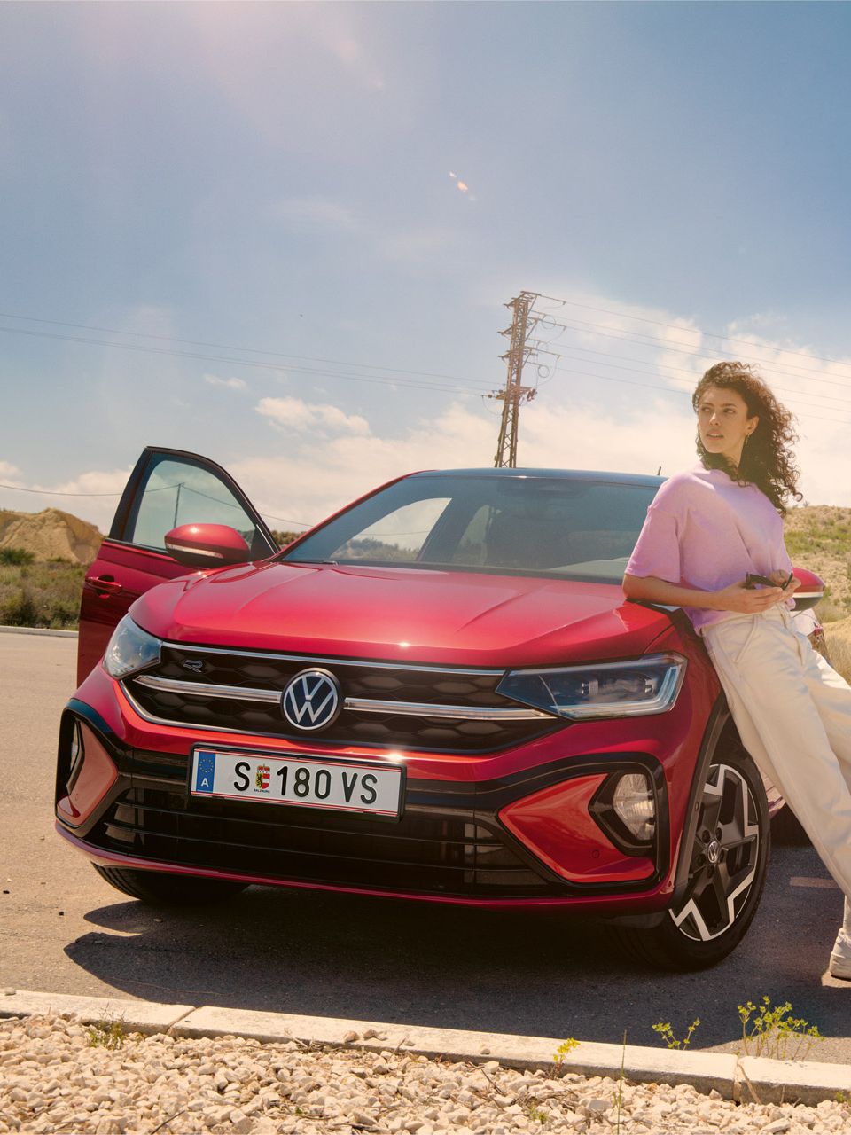 Frau lehnt an der Motorhaube eines VW Taigo auf einem Parkplatz in der Wüste. Mann fährt auf Skateboard dahinter entlang.
