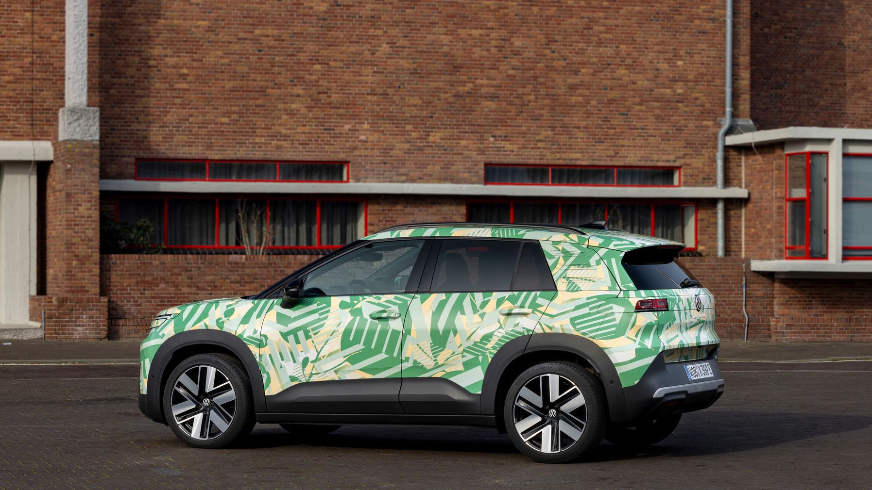 Ein grünes SUV mit tropischem Blattmuster steht vor einem roten Backsteingebäude mit mehreren Fenstern. Die Reifen sind modern und das Auto ist auf einer asphaltierten Fläche geparkt.