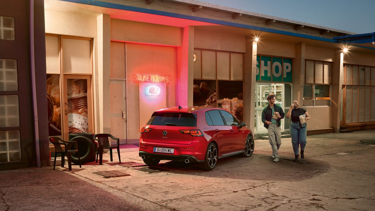 Roter VW Golf GTI steht abends vor einem Supermarkt, zwei junge Leute gehen mit Einkäufen daran vorbei.
