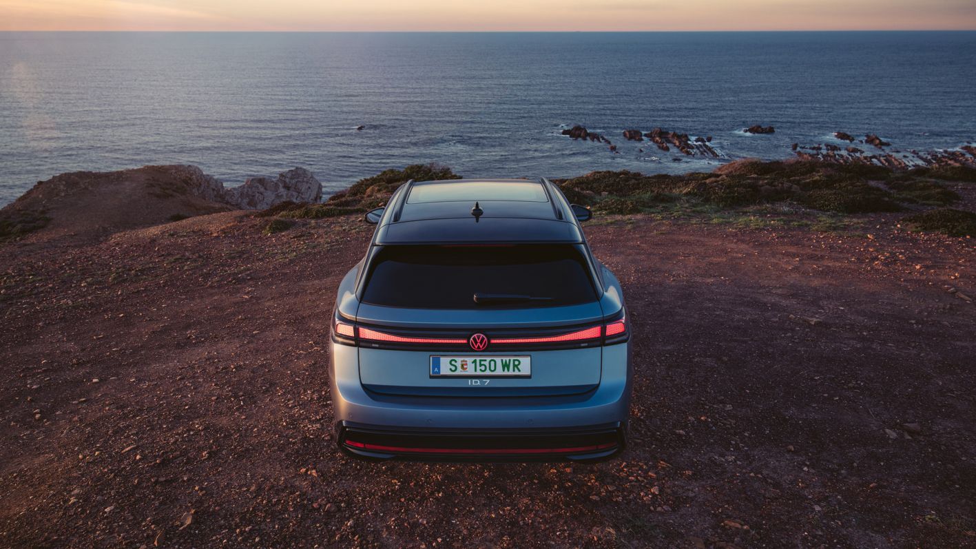 Ein blauer VW ID.7 Tourer in Heckansicht steht bei einer Klippe am Meer, die Heck-Lichtsignatur ist angeschaltet.