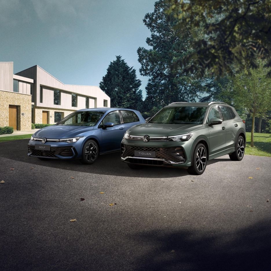 Zwei VW Modell, ein blauer Golf und ein grüner Tiguan, stehen nebeneinander auf einer Einfahrt vor einem Reihenhaus. Das Umfeld ist grün und bewaldet, das Wetter scheint klar zu sein.