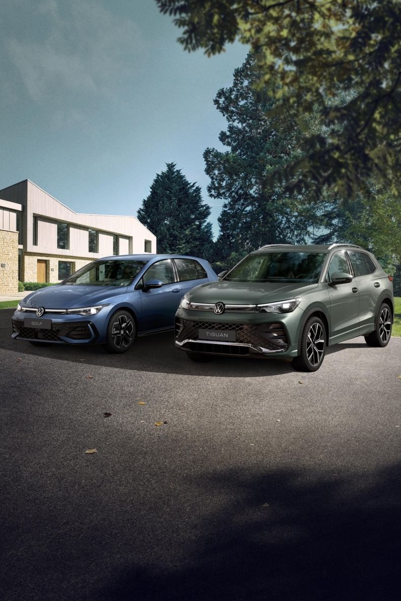 Zwei VW Modell, ein blauer Golf und ein grüner Tiguan, stehen nebeneinander auf einer Einfahrt vor einem Reihenhaus. Das Umfeld ist grün und bewaldet, das Wetter scheint klar zu sein.