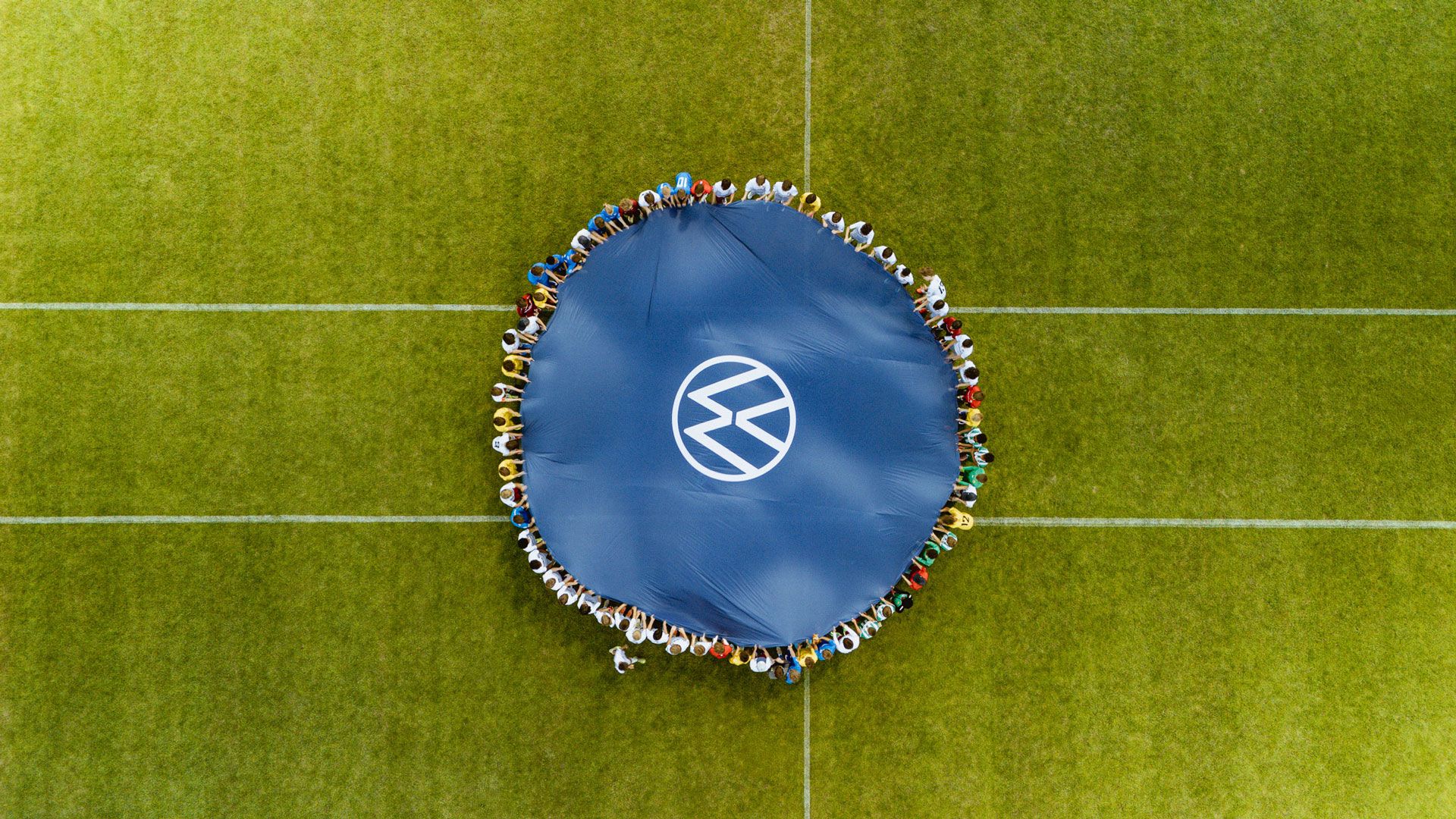Luftaufnahme eines Fußballfeldes, auf dessen Mittelpunkt ein großes, rundes blaues Tuch mit dem VW-Logo ausgebreitet ist. Rund um das Tuch stehen viele Menschen Schulter an Schulter im Kreis auf dem grünen Rasen.