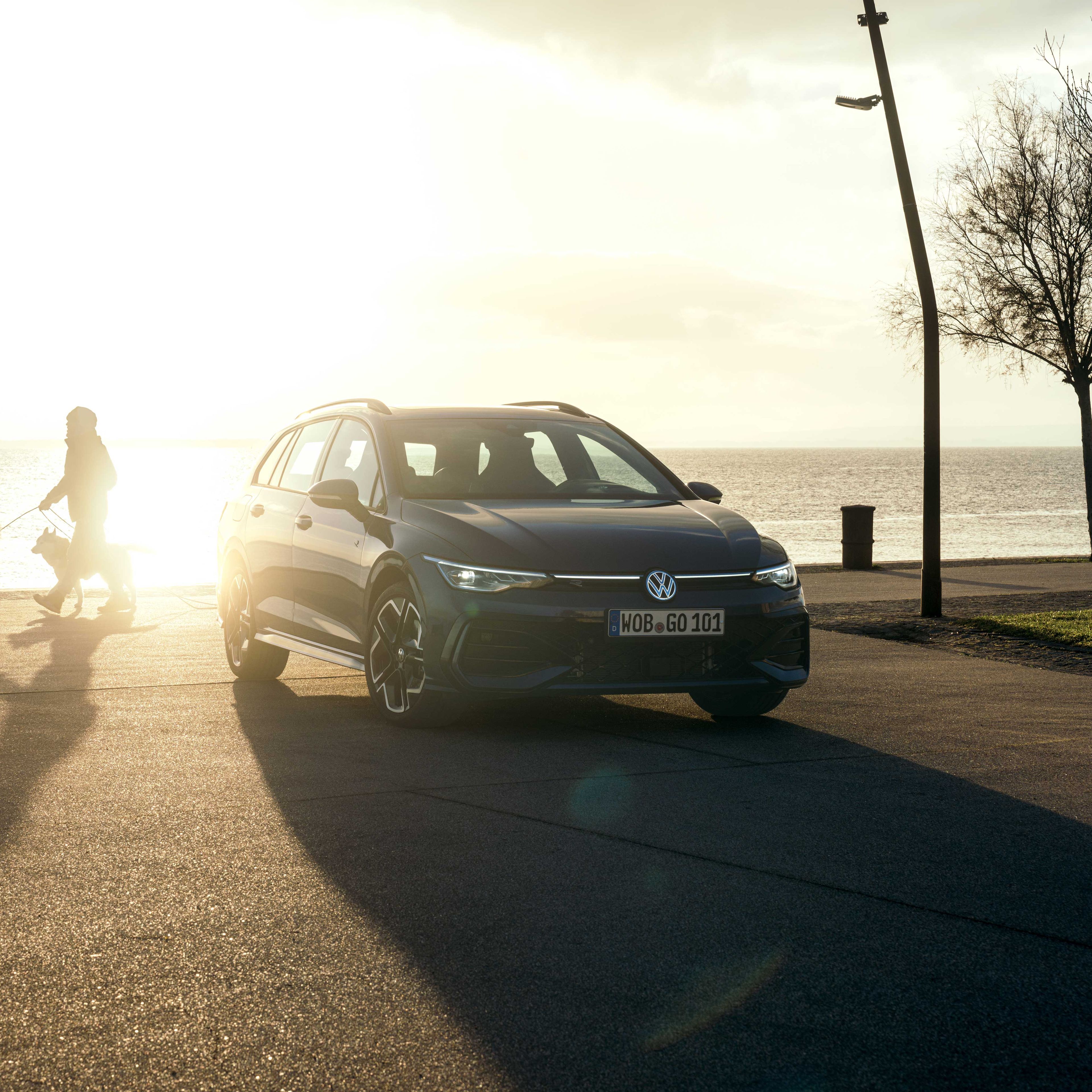 Ein dunkler Volkswagen Golf R-Line Kombi ist bei Sonnenaufgang an einer Küste geparkt. Eine Person geht mit zwei Hunden auf der Promenade spazieren.