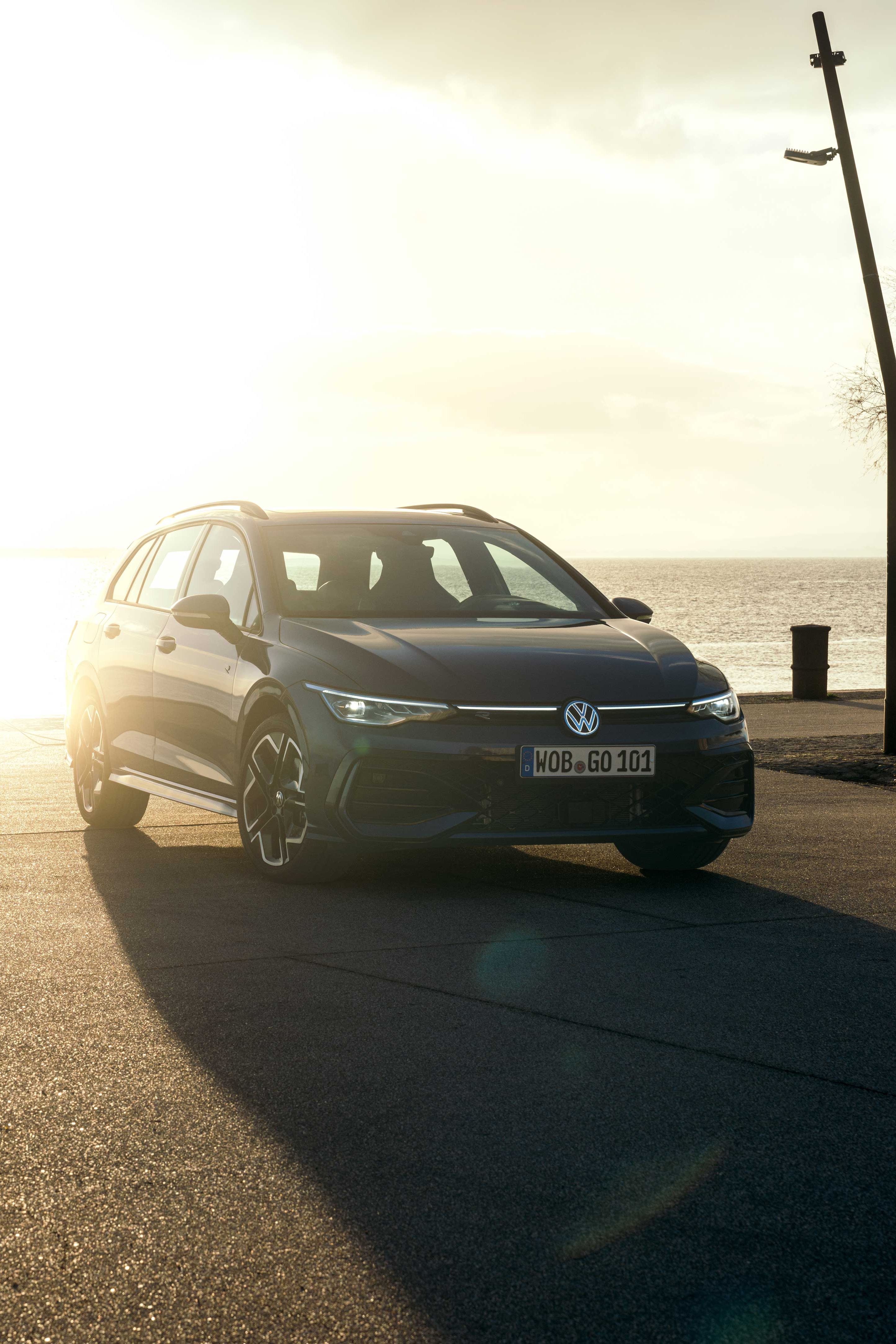 Ein dunkler Volkswagen Golf R-Line Kombi ist bei Sonnenaufgang an einer Küste geparkt. Eine Person geht mit zwei Hunden auf der Promenade spazieren.