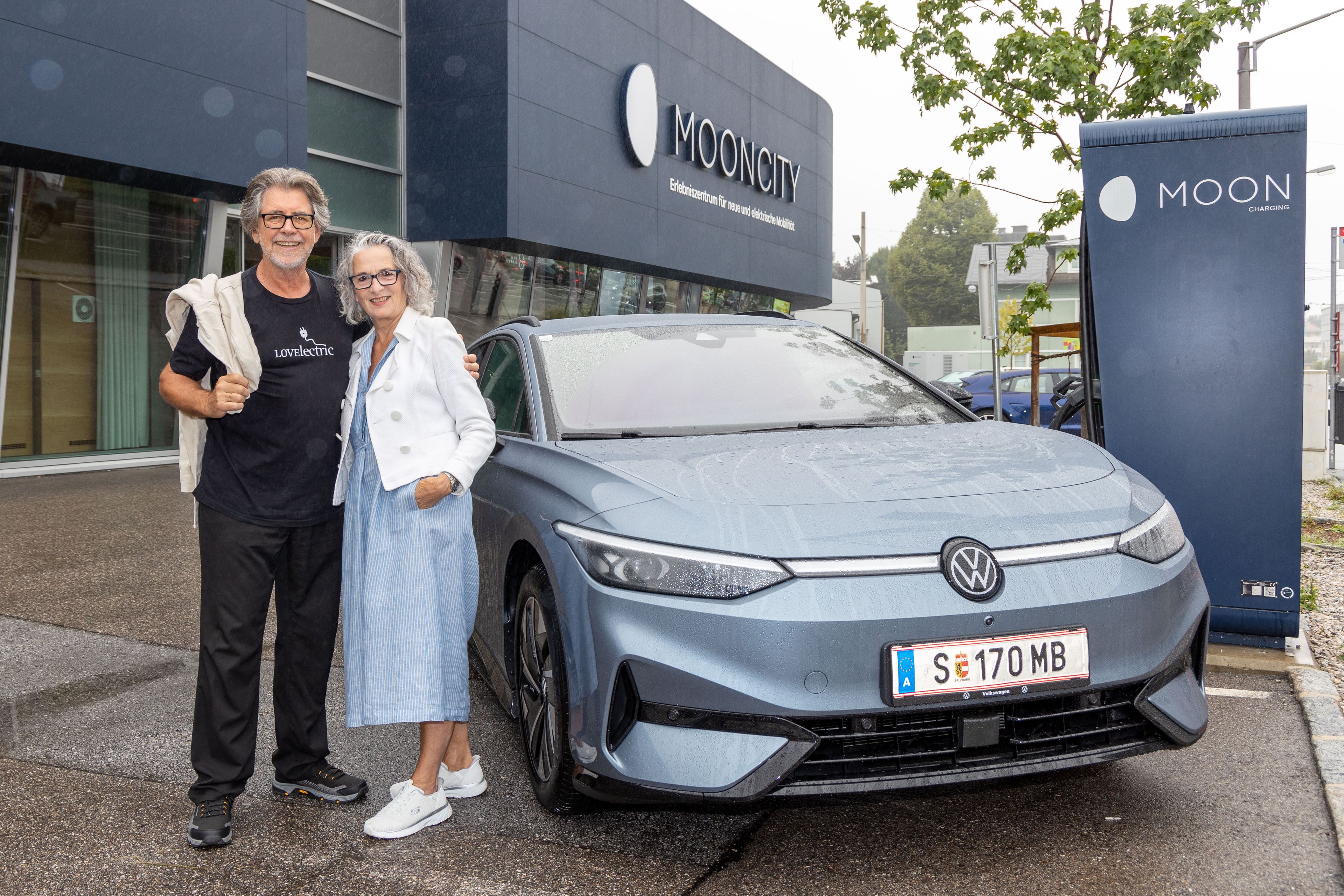 Ein Mann und eine Frau stehen vor einem ID.7 Tourer und lächeln. Im Hintergrund ist das Gebäude von Mooncity zu sehen, einem Ort für Elektromobilität. Es regnet leicht.