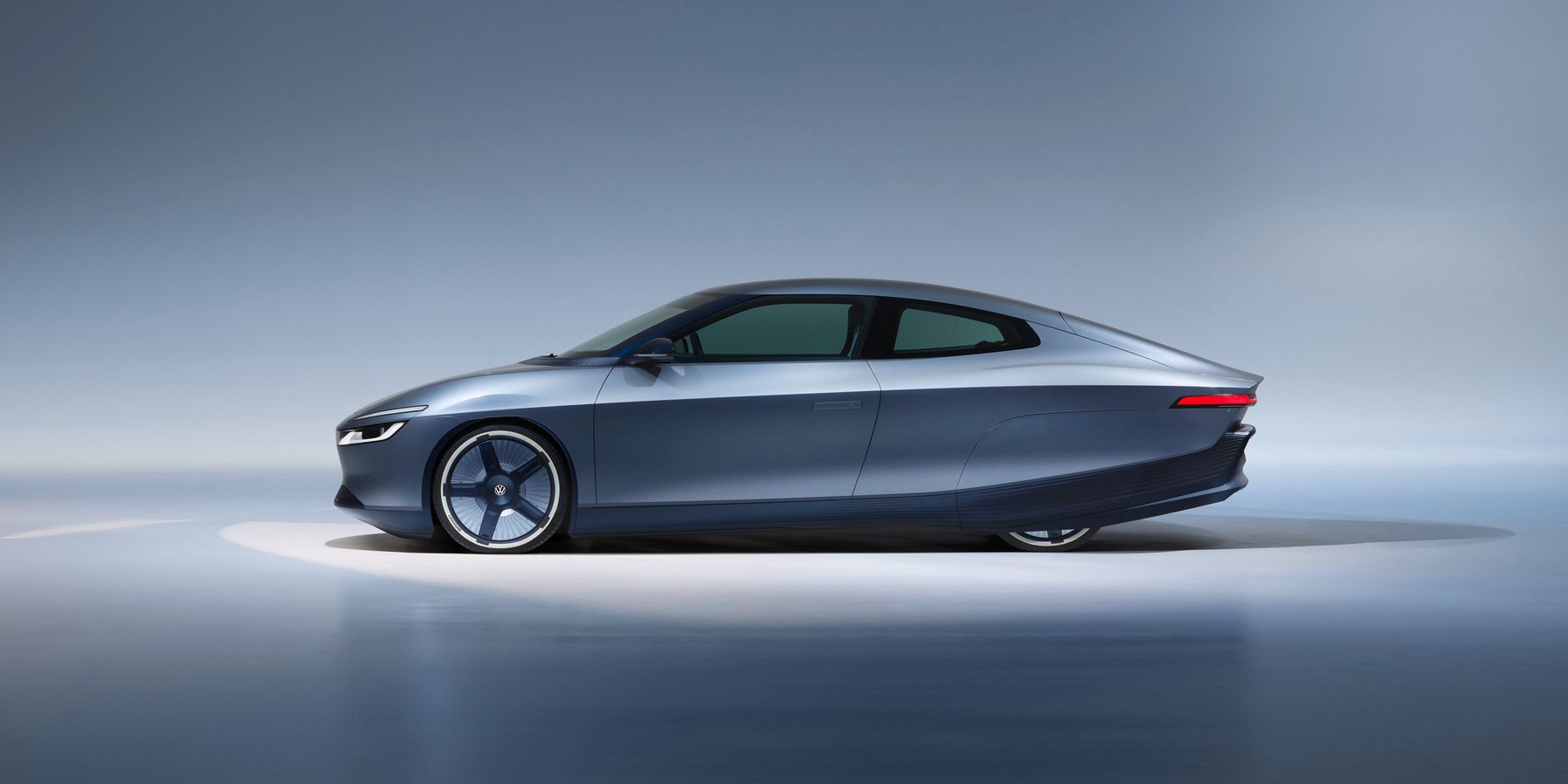 Seitlich aufgenommenes, futuristisches Elektroauto in dunkelgrauer Metallic-Lackierung vor neutralem, blau-grauem Studiohintergrund. Die Karosserie ist sehr flach und aerodynamisch mit glatten Flächen, verdeckten Türgriffen und schmalen Leuchten. Die Felgen haben ein nahezu geschlossenes, minimalistisches Design.