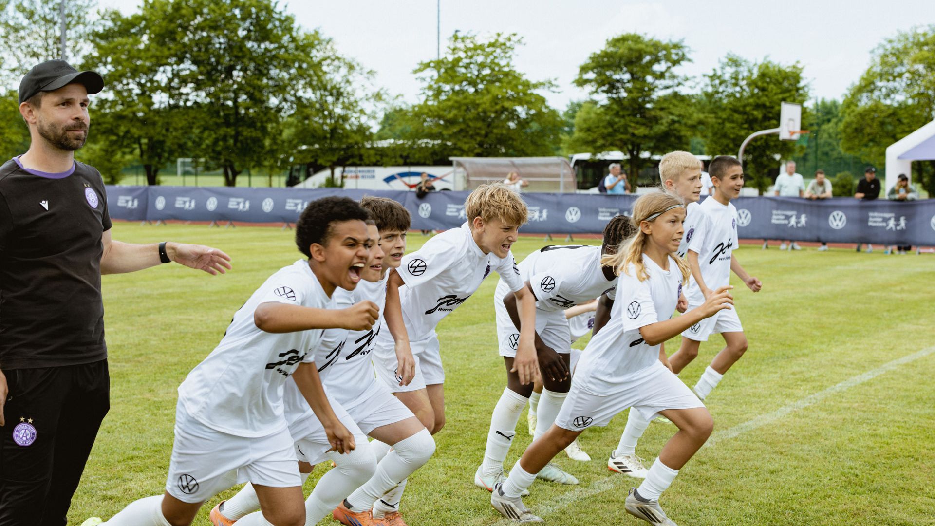 Eine Gruppe von Kindern in weißen Fußballtrikots sprintet nebeneinander über ein Spielfeld. Ein Trainer in dunkler Kleidung steht am Rand und beobachtet sie. Im Hintergrund sind Bäume, Werbebanner und weitere Sporteinrichtungen zu sehen.