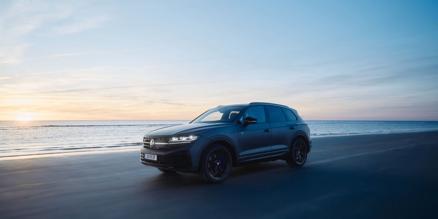 Ein dunkler Touareg R fährt bei Sonnenuntergang auf einem Strand neben dem Meer. Der Himmel ist überwiegend blau, mit einigen orangefarbenen und gelben Tönen der untergehenden Sonne. Das Meer ist ruhig im Hintergrund.