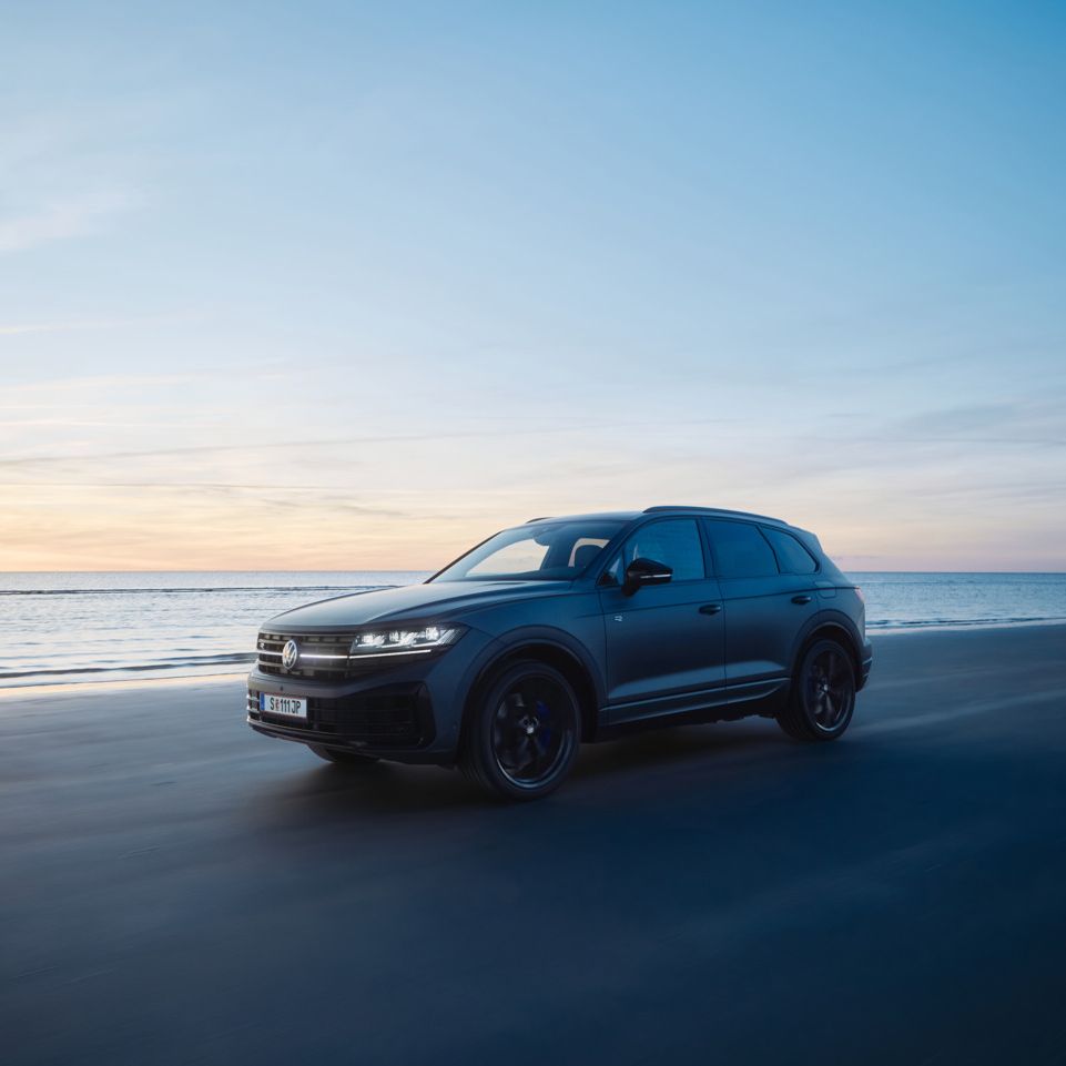 Ein dunkler Touareg R fährt bei Sonnenuntergang auf einem Strand neben dem Meer. Der Himmel ist überwiegend blau, mit einigen orangefarbenen und gelben Tönen der untergehenden Sonne. Das Meer ist ruhig im Hintergrund.