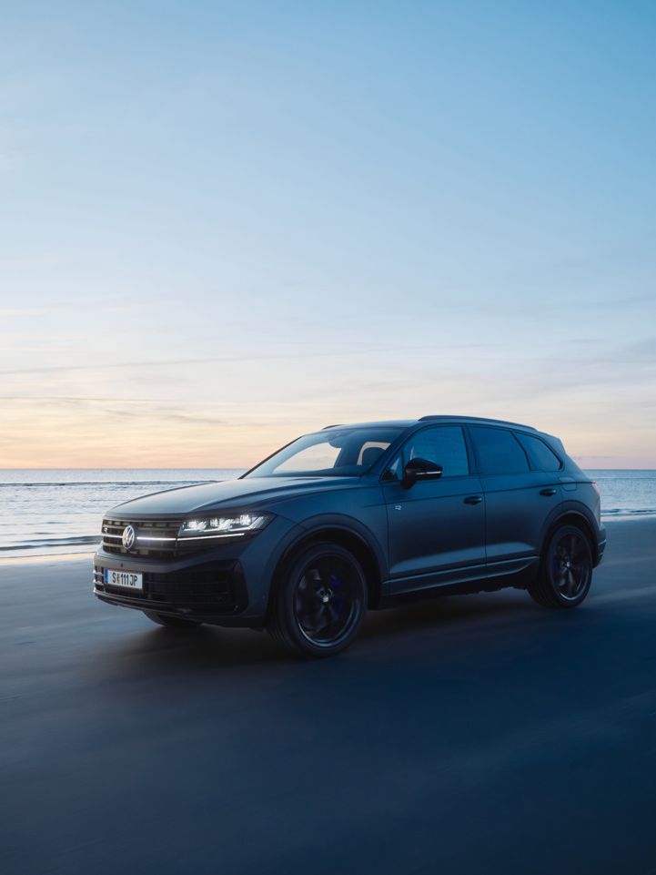 Ein dunkler Touareg R fährt bei Sonnenuntergang auf einem Strand neben dem Meer. Der Himmel ist überwiegend blau, mit einigen orangefarbenen und gelben Tönen der untergehenden Sonne. Das Meer ist ruhig im Hintergrund.