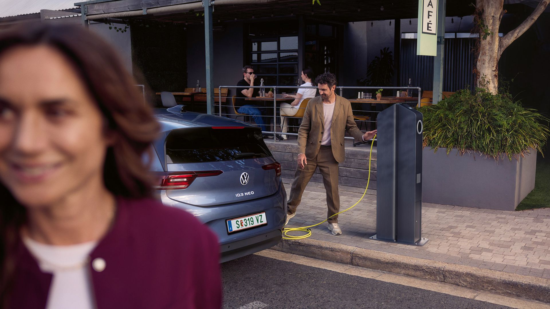 Eine Frau geht auf einer Straße. Hinter ihr läd ein Mann einen VW ID.3 Neo an einer Ladesäule vor einem Café