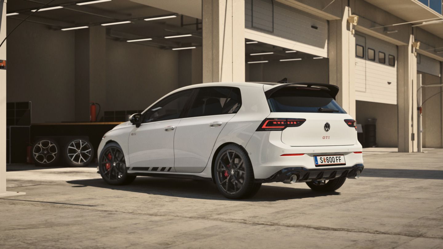 Ein weißer Volkswagen Golf GTI Clubsport steht in einer Werkstatt oder Garage. Das Auto hat schwarze Felgen und Details, sowie ein österreichisches Kennzeichen. Im Hintergrund sind Räder und Werkstattausrüstung zu sehen.