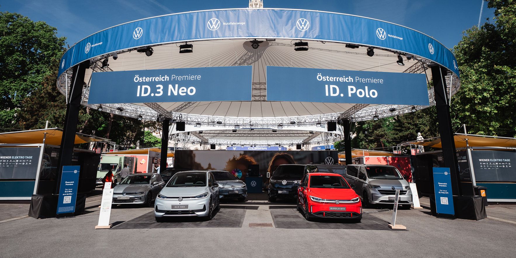 Offener VW-Ausstellungsstand im Freien mit mehreren Elektro- und Hybridfahrzeugen, im Vordergrund ein silberner, ein grauer, ein roter und ein weiterer silberner Wagen. Über dem Stand hängen große blaue Banner mit der Aufschrift „Österreich Premiere ID.3 Neo“ und „Österreich Premiere ID. Polo“. Im Hintergrund sind Bäume, Infostände und ein historisches Gebäude mit Turm zu sehen.