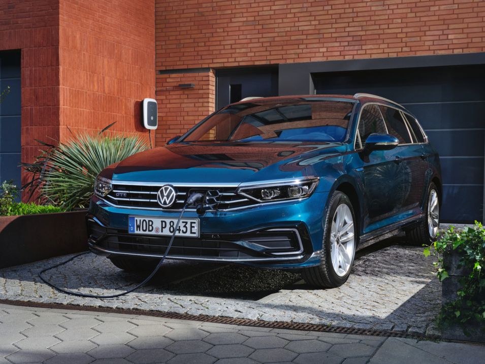 Ein blauer Volkswagen Passat GTE ist an einer Ladestation in einer modernen Wohngegend angeschlossen. Das Fahrzeug steht auf einer gepflasterten Einfahrt vor einem Gebäude mit roten Ziegeln. Pflanzen sind um die Einfahrt herum angeordnet.