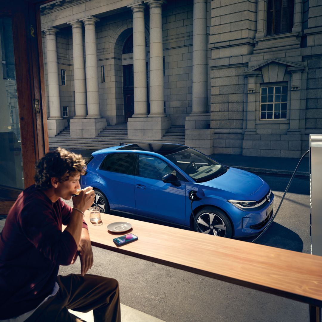 Ein Mann sitzt in einem Café, durch ein offenes Fenster ist der blaue VW ID. Polo Style zu sehen, der an einer Ladestation lädt.