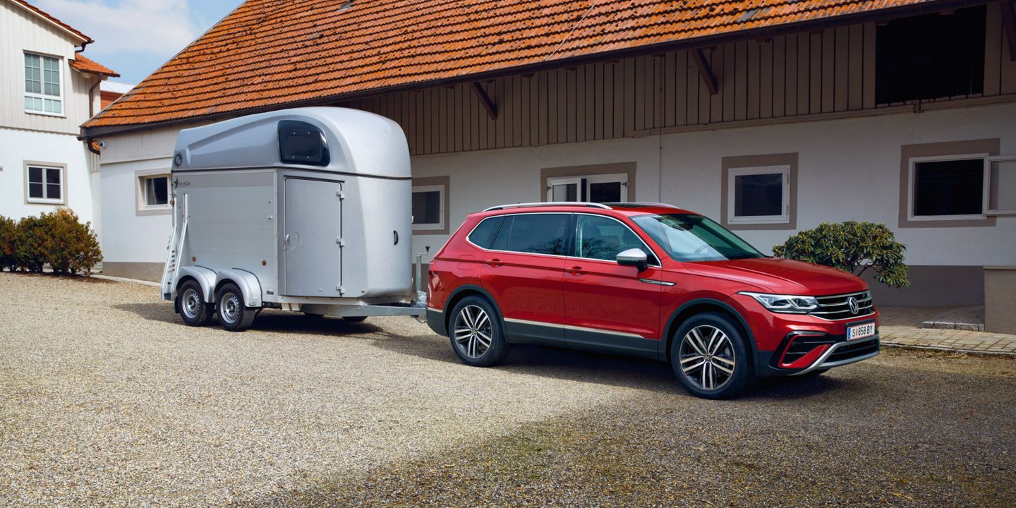 Roter Tiguan Allspace mit silbernem Pferdeanhänger steht auf einem Kieshof vor einem langgezogenen Wohn- oder Wirtschaftsgebäude mit roten Dachziegeln. Das Fahrzeug ist seitlich zu sehen, der Anhänger ist zweiachsig und geschlossen. Im Hintergrund befinden sich weiße Hauswände mit mehreren Fenstern und Türen.