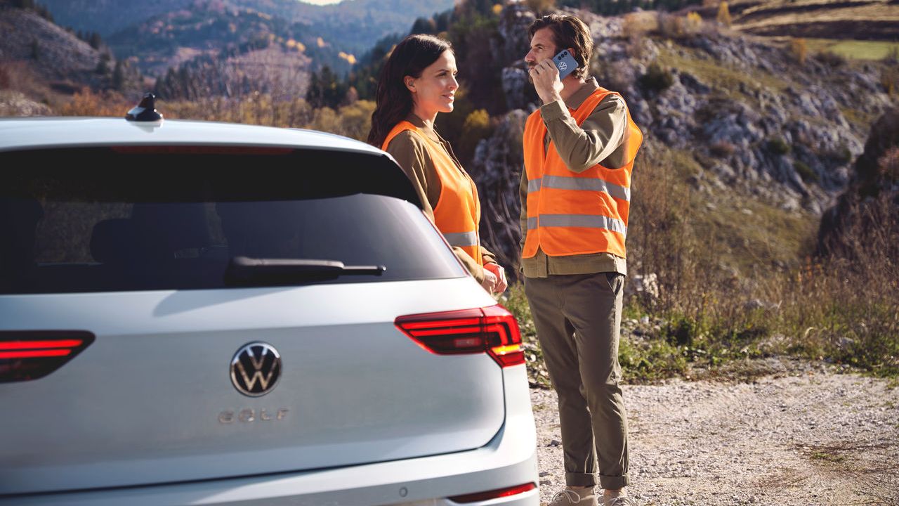 Zwei Personen in orangefarbenen Sicherheitswesten stehen vor einem Volkswagen Golf. Eine der Personen telefoniert.