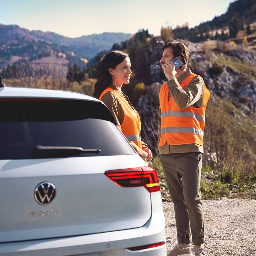 Zwei Personen in orangefarbenen Sicherheitswesten stehen vor einem Volkswagen Golf. Eine der Personen telefoniert.