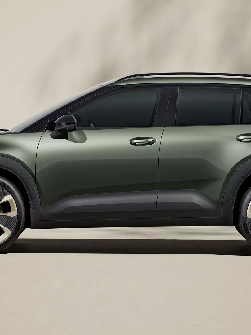 Ein modernes SUV in dunklem Grün steht vor einem neutralen Hintergrund. Das Fahrzeug hat große Felgen und eine markante Silhouette. Schattenmuster sind auf dem Hintergrund zu erkennen.
