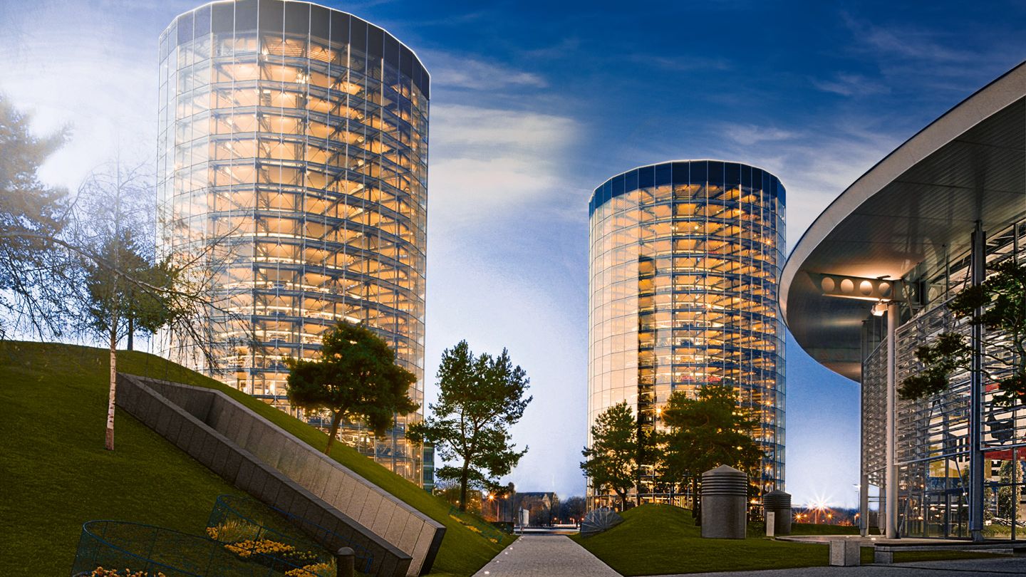 Panorama der Autostadt in Wolfsburg bei Abenddämmerung mit zwei runden, voll verglasten Hochgaragen, in denen zahlreiche beleuchtete Fahrzeugebenen zu sehen sind. Rechts steht ein futuristisches, halbkreisförmiges Gebäude mit Glasfassade, links und in der Mitte verlaufen Wege und begrünte Hügel mit einzelnen Bäumen. Die erleuchteten Gebäude spiegeln sich in den Glasflächen und vermitteln eine high-tech, urbane Atmosphäre.