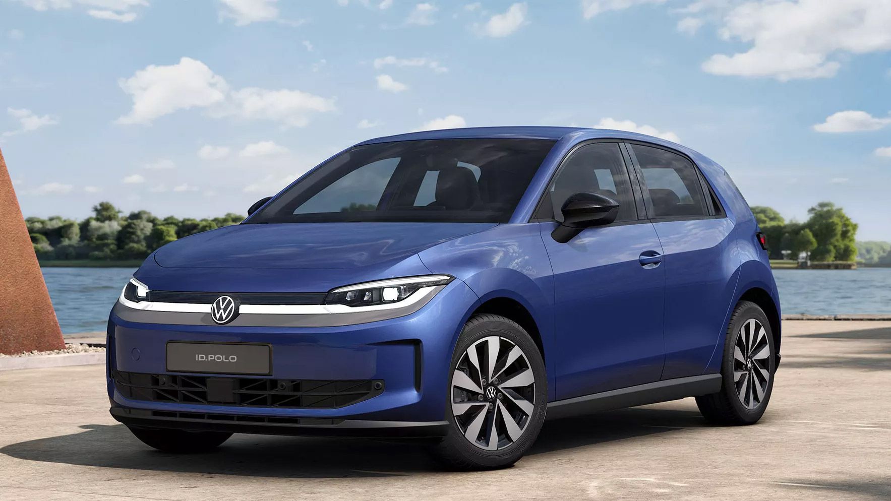 Ein blauer VW ID. Polo Style von schräg vorne steht auf einem hellen Weg vor einem See.