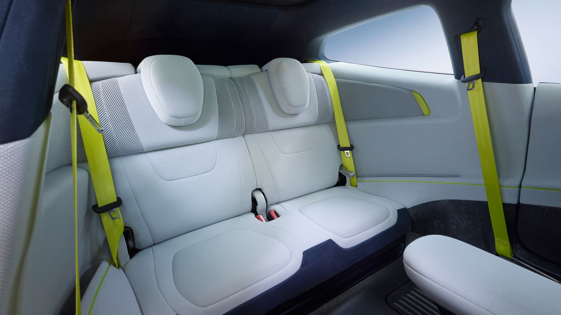 Hintere Sitzreihe in einem modernen Auto mit hellen, fast weißen Polstern und integrierten Kopfstützen. Auffällig sind die neongelben Sicherheitsgurte an den Seiten, die einen starken Kontrast zum ansonsten minimalistischen, grau-weißen Innenraum bilden. Die Gestaltung wirkt geräumig, futuristisch und aufgeräumt, mit klaren Linien und glatten Oberflächen.
