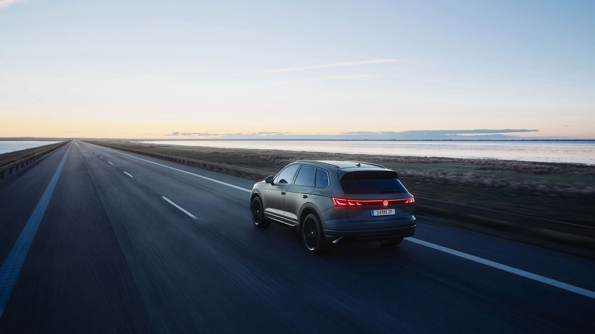 Ein dunkler Touareg R fährt auf einer leeren Straße entlang der Küste bei Sonnenuntergang. Die Straße erstreckt sich in die Ferne, während der Himmel im Hintergrund mit warmen Farben erleuchtet ist. Links und rechts der Straße befinden sich Wasserflächen und Grünflächen.