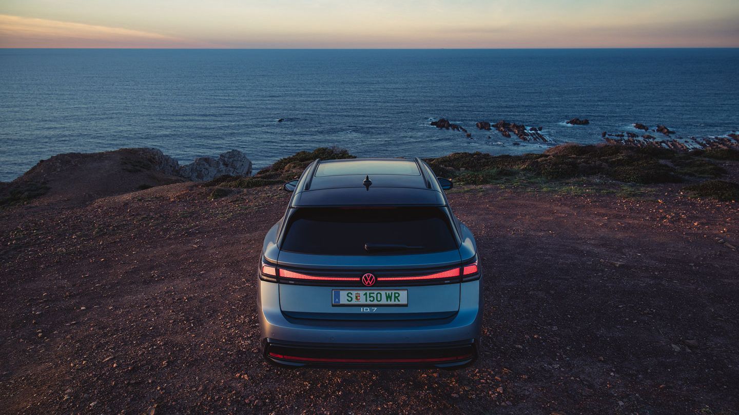 Blauer VW ID.7 Tourer in Heckansicht steht auf einer Klippe am Meer, die Heck-Lichtsignatur ist angeschaltet.