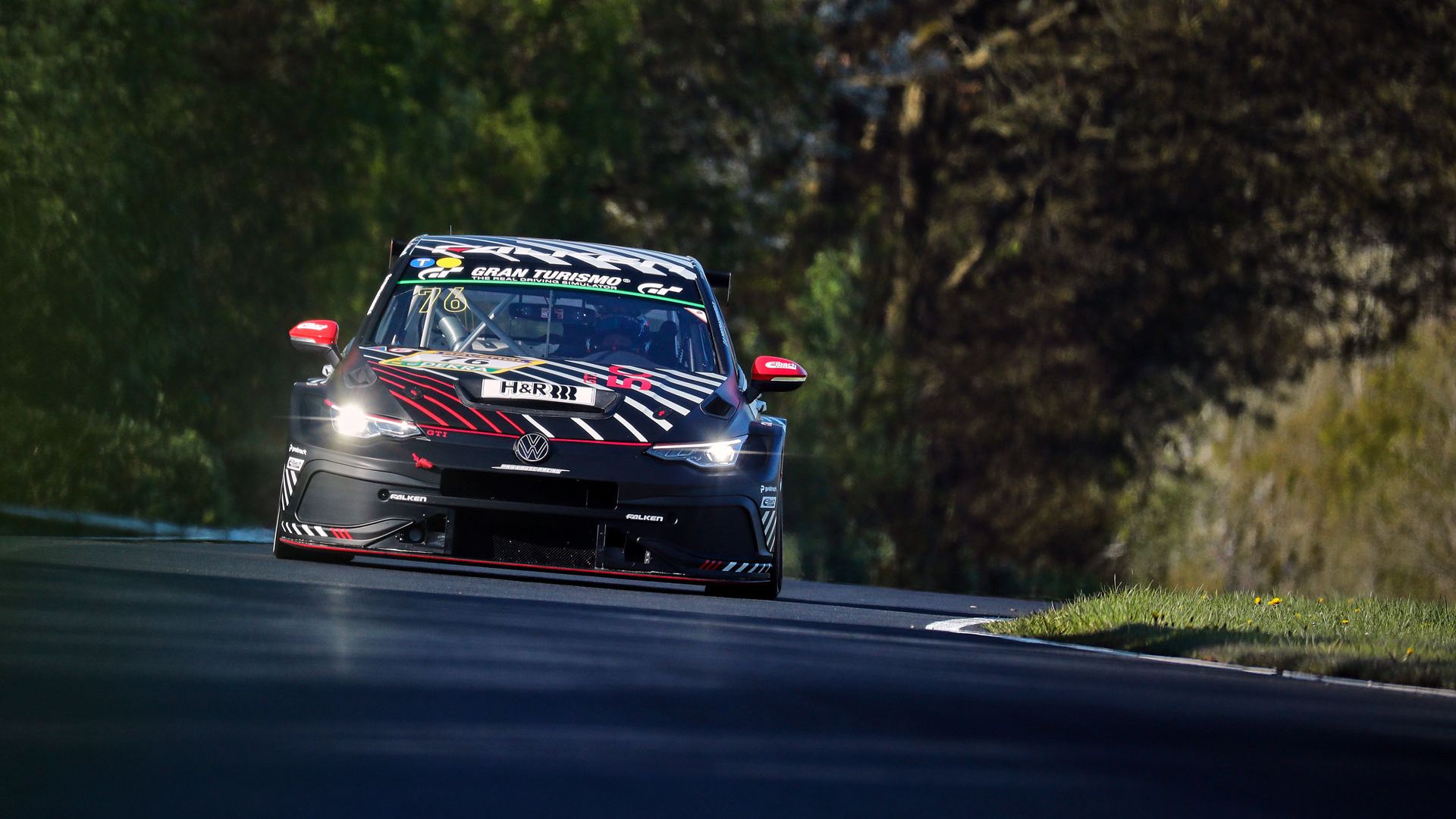 Ein Golf GTI Clubsport fährt auf einer Rennstrecke durch eine landschaftlich grüne Umgebung. Die Straße führt um eine Kurve und der Wagen hat auffällige rote und schwarze Markierungen. Der Hintergrund zeigt Bäume und Vegetation.