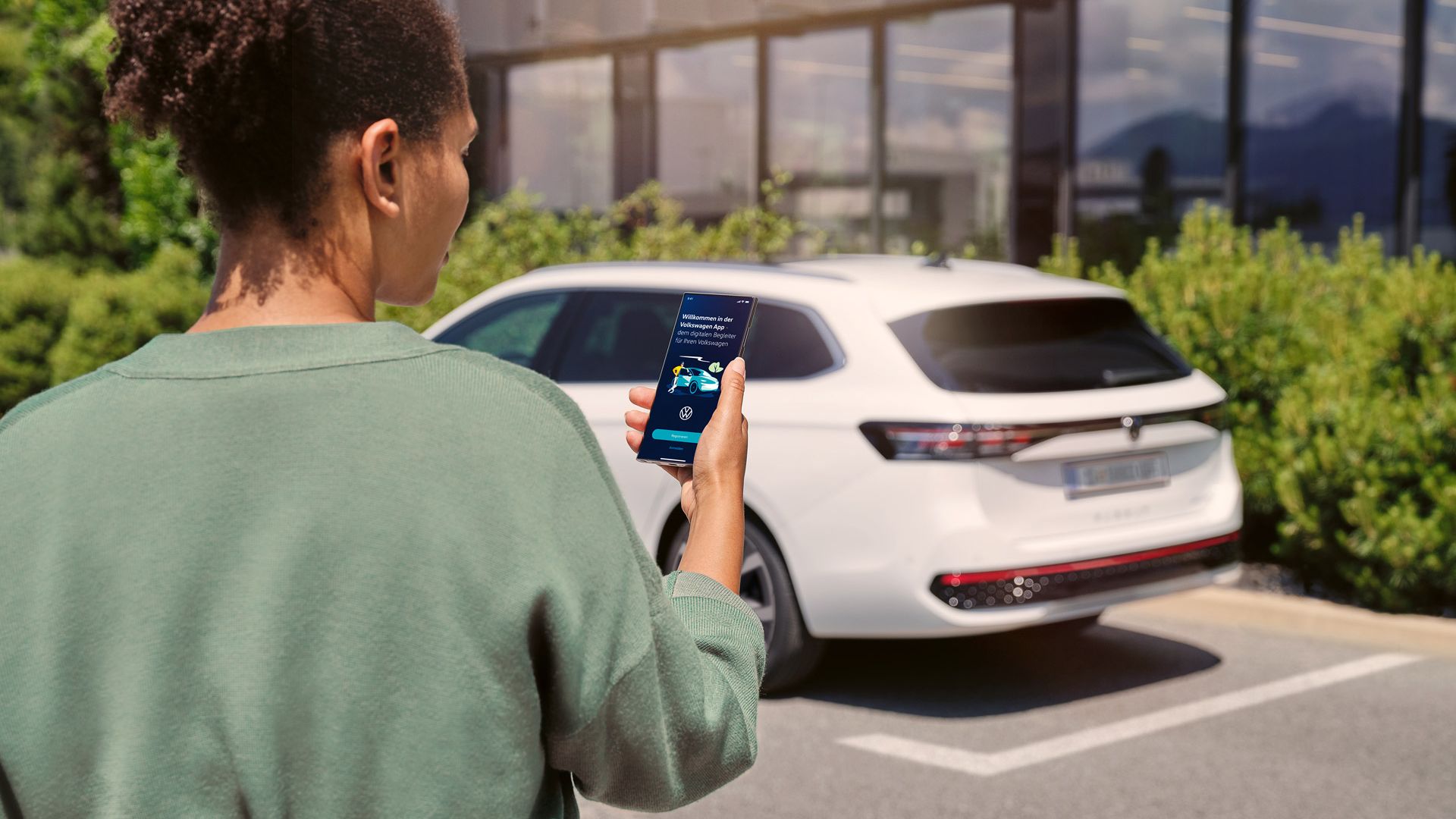  Ein weißer VW Passat parkt seitlich vor einem Gebäude. Eine Frau in Rückenansicht schaut auf ihr Smartphone.