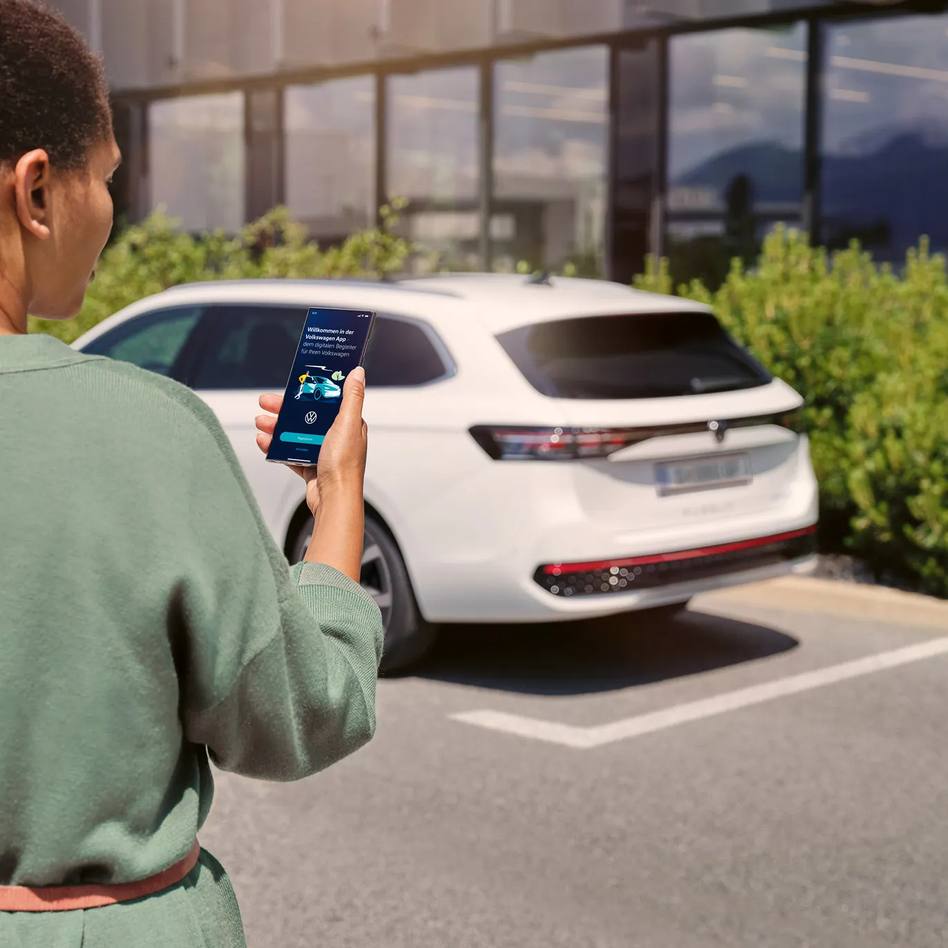  Ein weißer VW Passat parkt seitlich vor einem Gebäude. Eine Frau in Rückenansicht schaut auf ihr Smartphone.