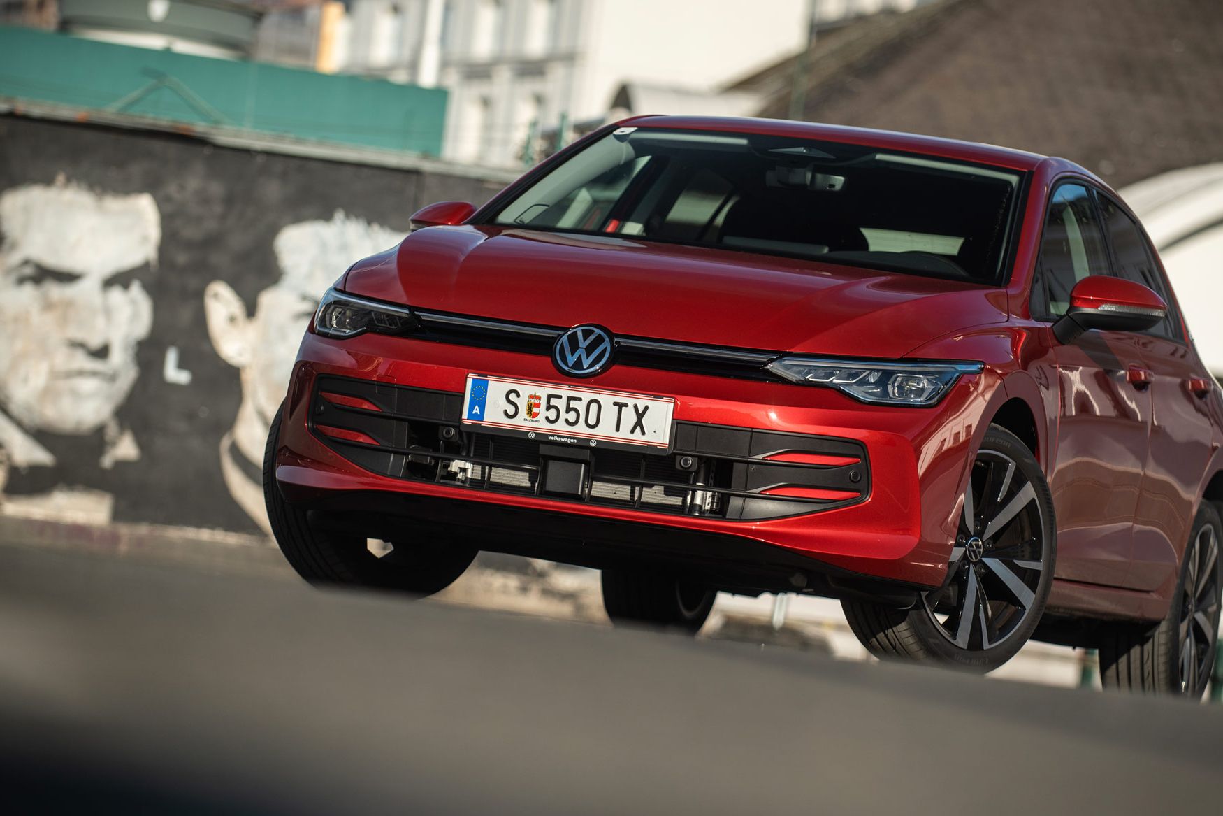 Roter Volkswagen Golf, schräg von vorne fotografiert, steht auf einer Straße vor einer Häuserkulisse.  
Das Auto wirkt dynamisch, als wäre es in Bewegung, und die Sonne erzeugt Reflexionen auf dem Lack.