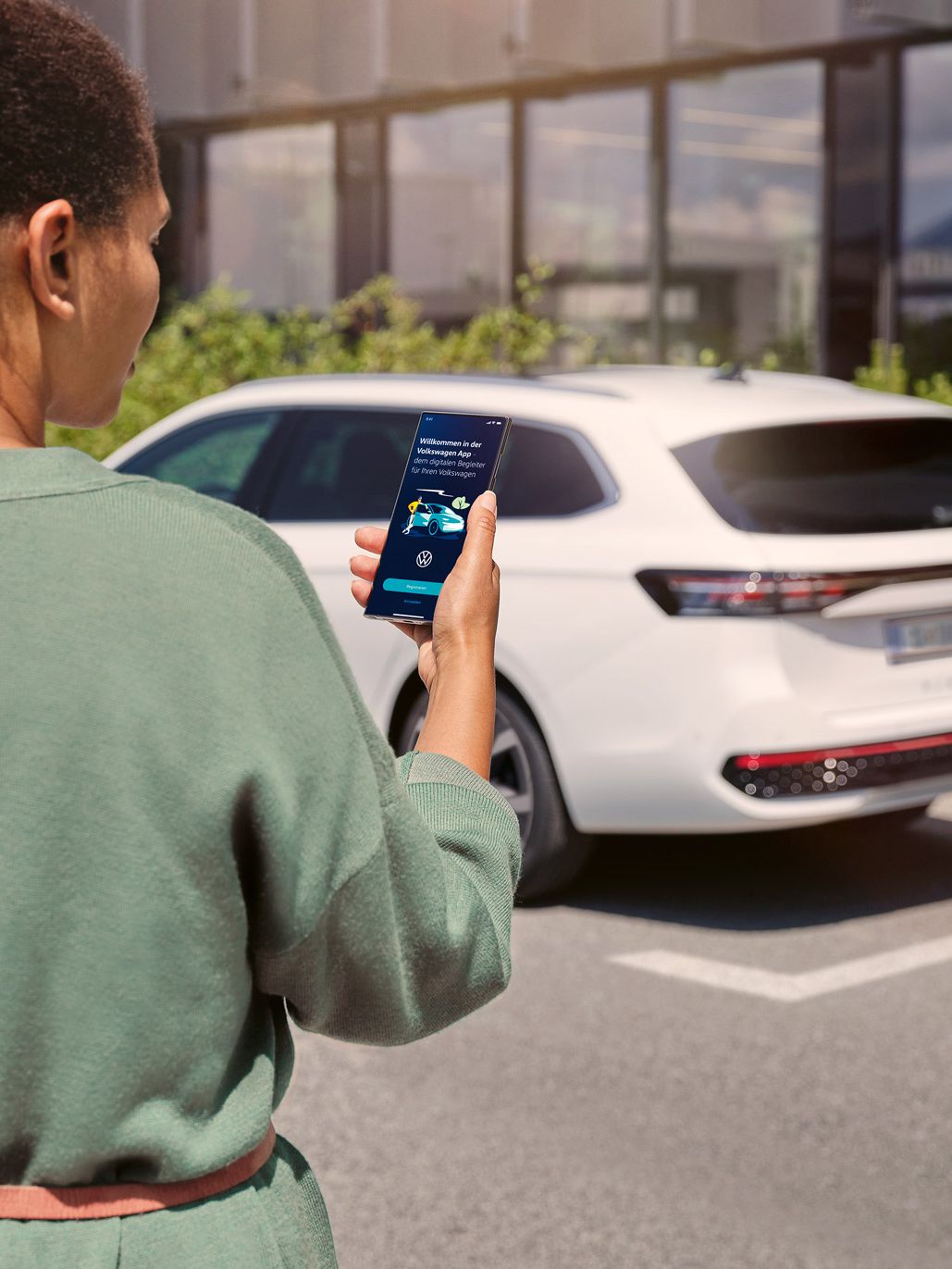 Ein weißer VW Passat parkt seitlich vor einem Gebäude. Eine Frau in Rückenansicht schaut auf ihr Smartphone.
