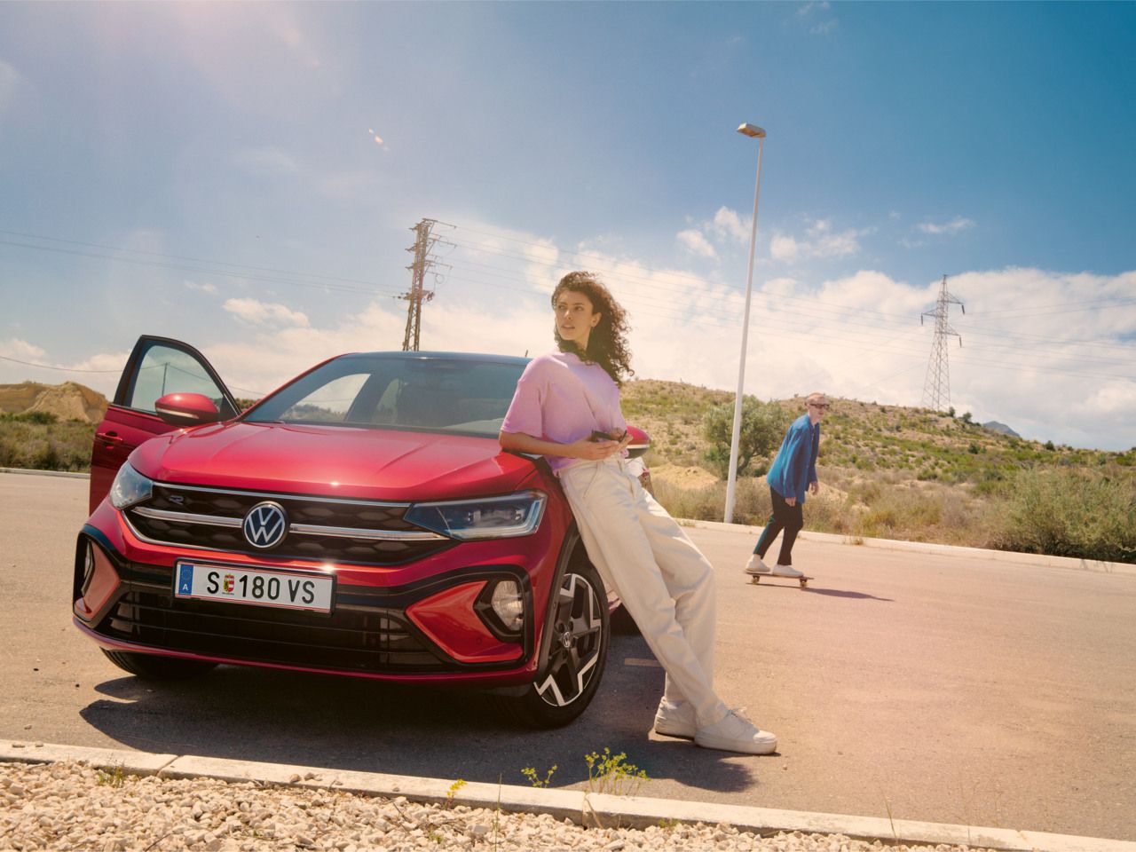Eine Frau lehnt an der Motorhaube eines VW Taigo auf einem Parkplatz. Hinter ihr fährt ein Mann auf einem Skateboard vorbei.