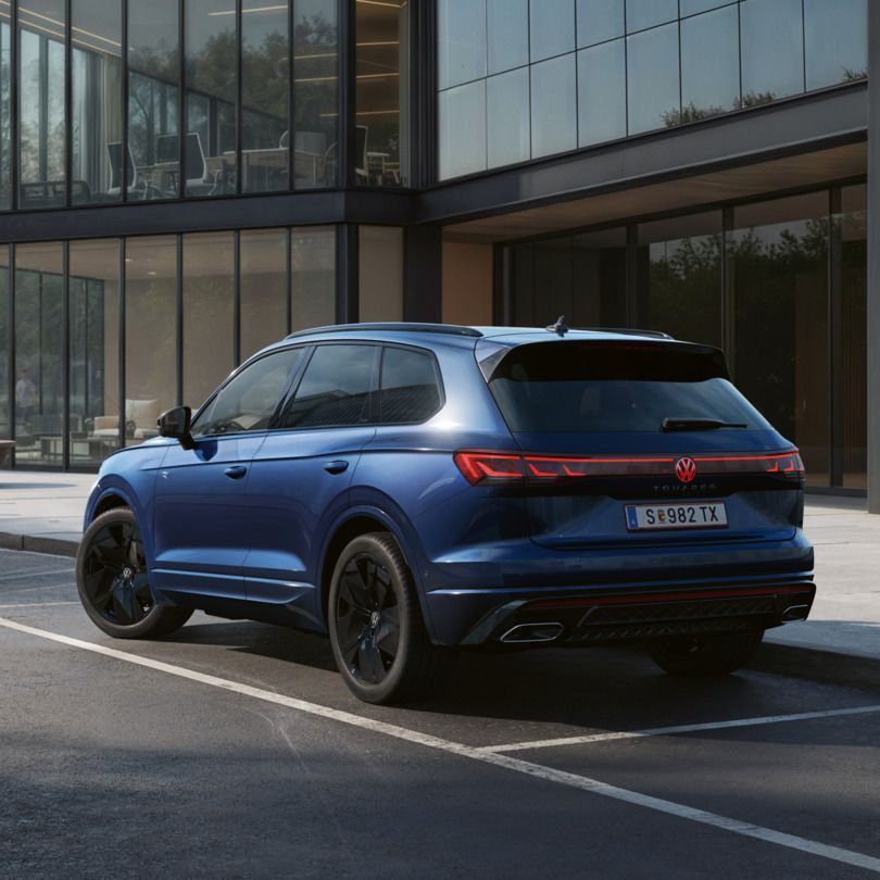 Ein blauer VW Touareg FINAL EDITION  steht vor einem Bürogebäude mit Glasfassade - Ansicht von schräg hinten.