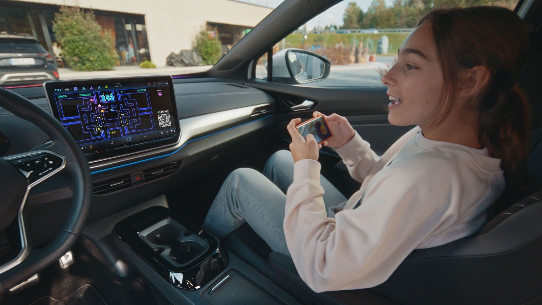 Eine Person sitzt entspannt im Fahrersitz eines Autos und spielt ein Videospiel auf ihrem Smartphone. Das Auto hat ein großes eingebautes Display auf dem Armaturenbrett, das ebenfalls ein Videospiel zeigt. Im Hintergrund ist die Außenseite eines Gebäudes zu sehen.