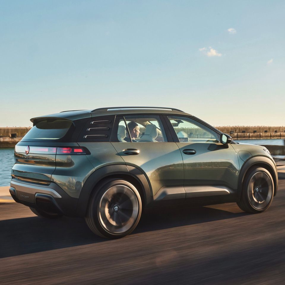 Ein grüner ID. Cross fährt auf einer Straße entlang der Küste. Im Hintergrund sind einige Flaggen und ein Hafengebiet zu sehen.