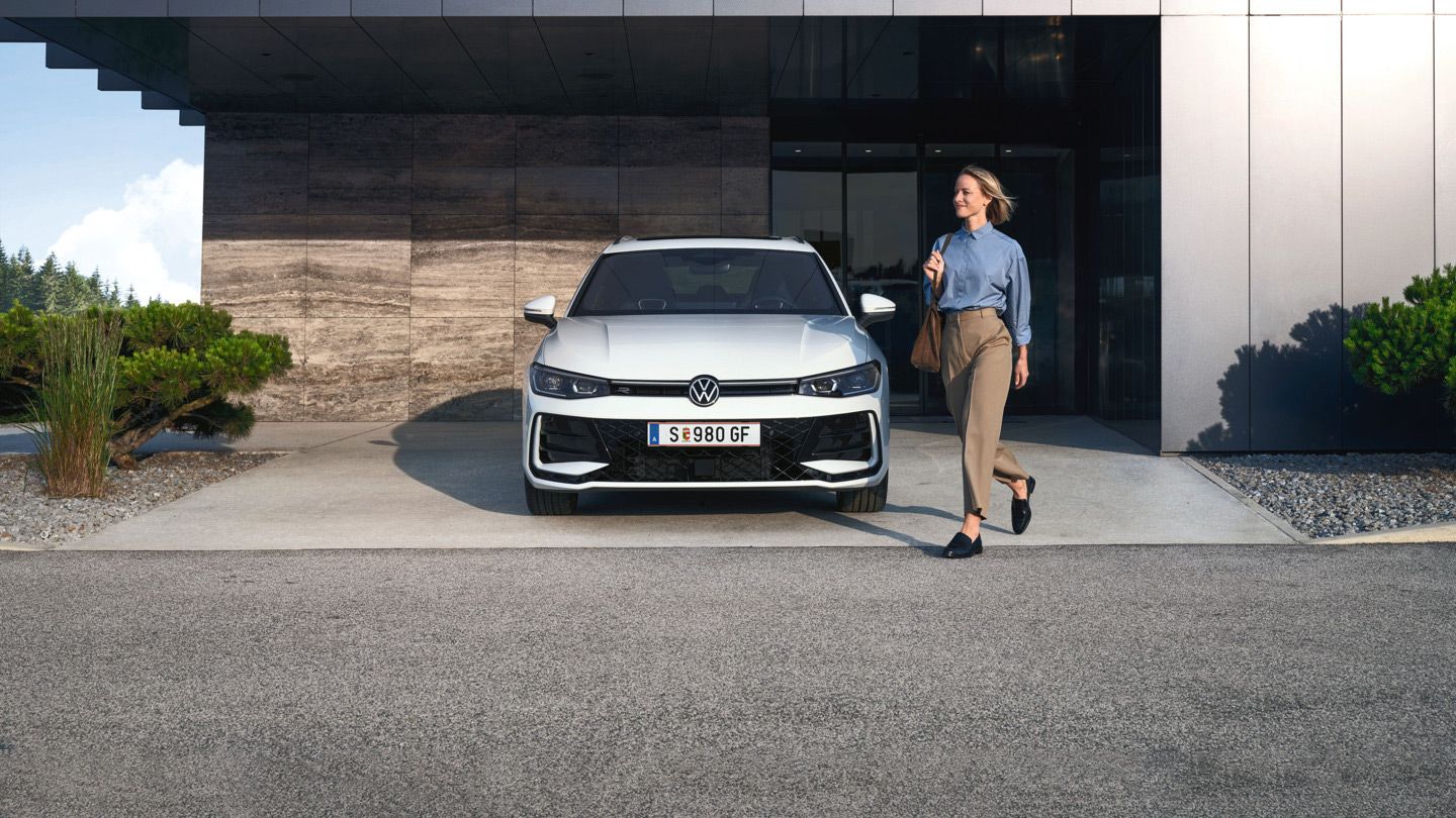 Frontansicht eines weißen VW Passat R-Line, eine Frau geht an ihm vorbei.