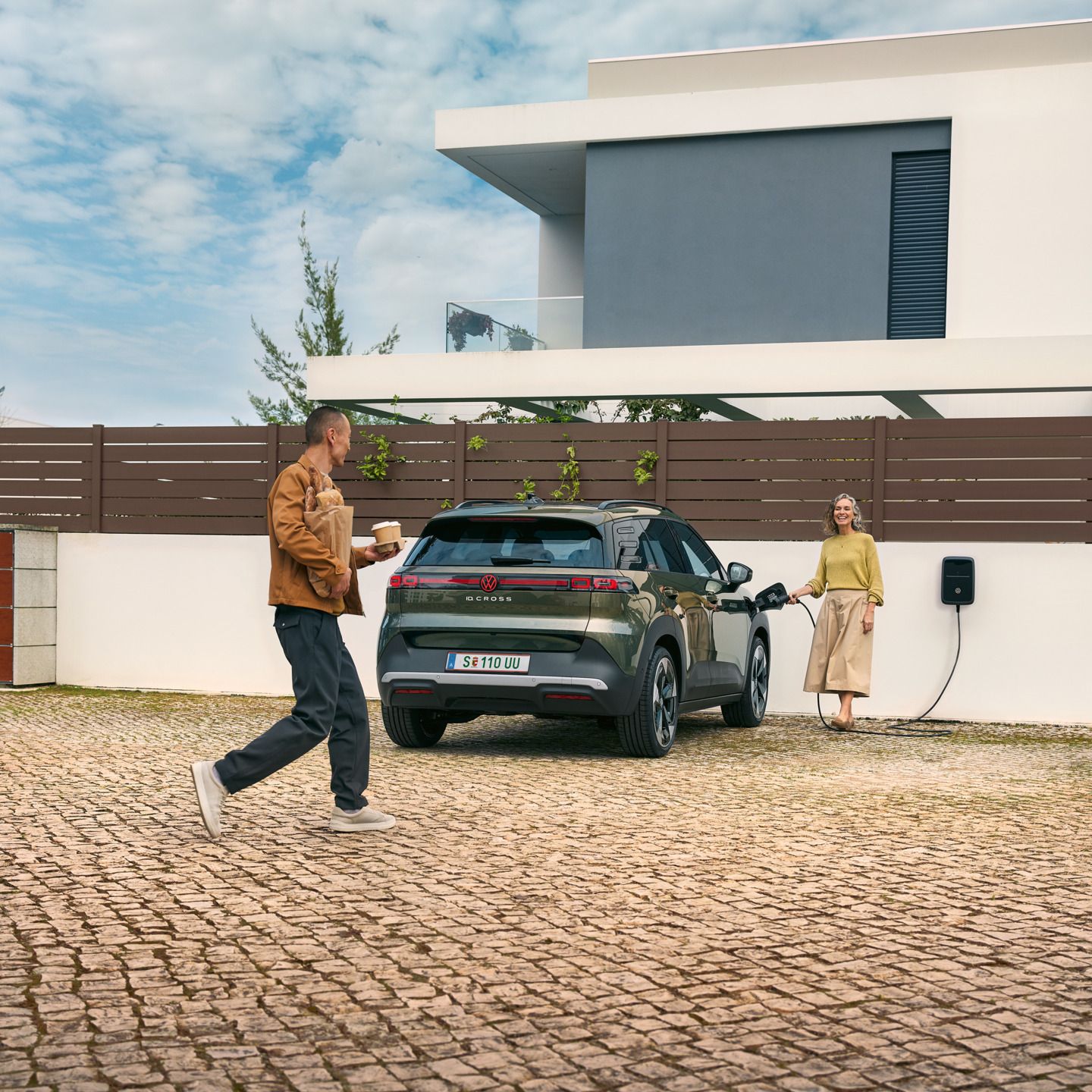 Eine Frau steckt ein Ladekabel einer Wallbox in einen grünen VW ID. Cross, ein Mann geht auf sie zu und hat Kaffeebecher in der Hand und Brot unter dem Arm.