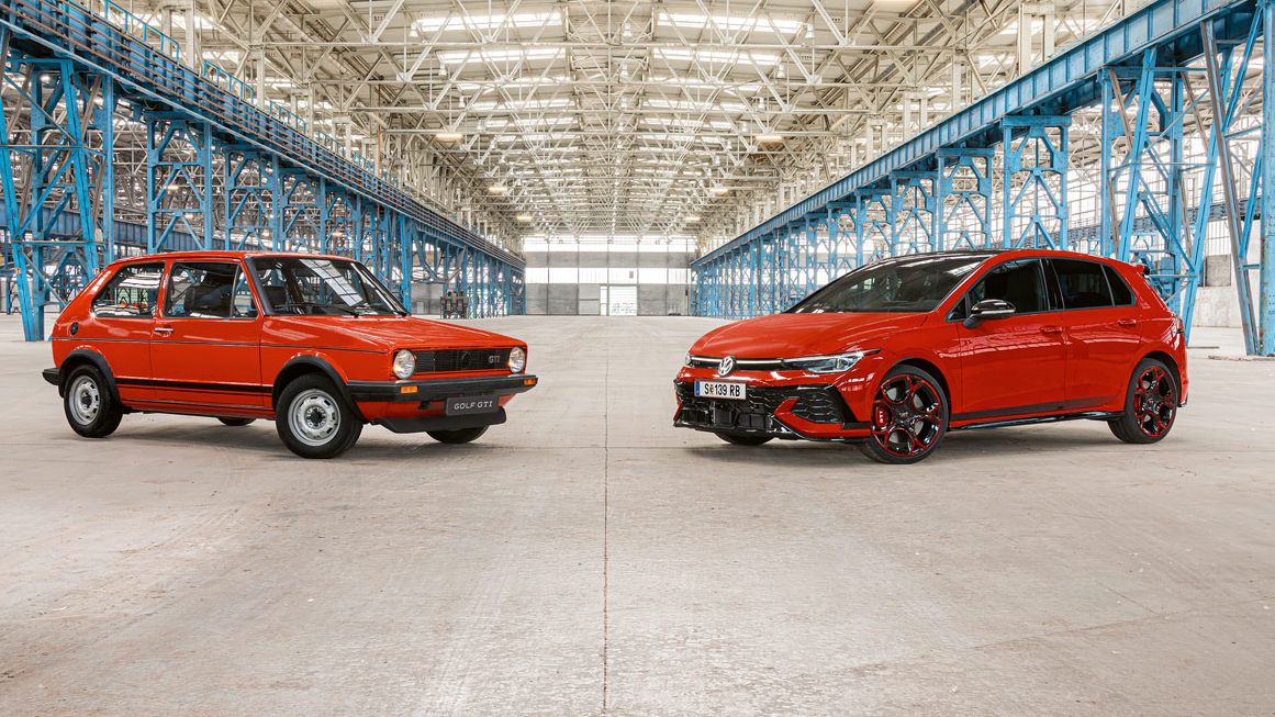 Zwei rote Golf GTI, das erste und das neuste Modell, stehen in einer großen Industriehalle mit vielen blauen Stahlträgern und einem hohen, hellen Dach.