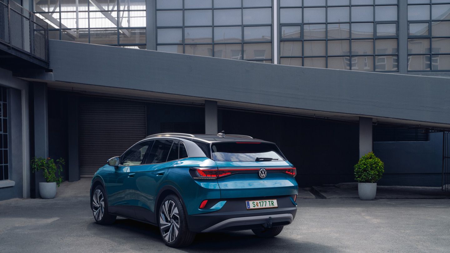 Ein blauer Volkswagen ID.4 steht geparkt in einem modernen, städtischen Umfeld. Das Fahrzeug zeigt seine Rückansicht. Im Hintergrund sind Gebäude aus Glas und Beton zu sehen.