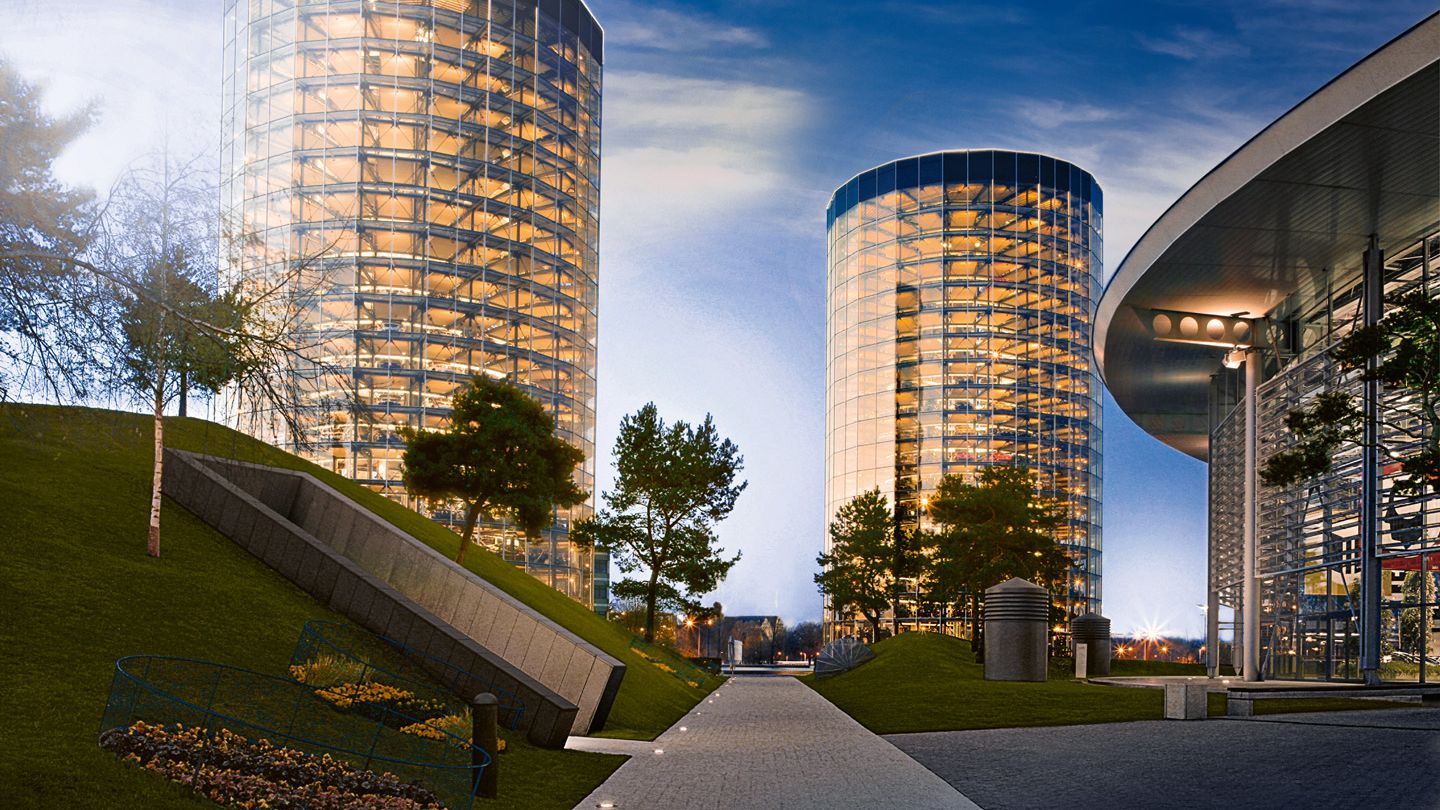 Panorama der Autostadt in Wolfsburg bei Abenddämmerung mit zwei runden, voll verglasten Hochgaragen, in denen zahlreiche beleuchtete Fahrzeugebenen zu sehen sind. Rechts steht ein futuristisches, halbkreisförmiges Gebäude mit Glasfassade, links und in der Mitte verlaufen Wege und begrünte Hügel mit einzelnen Bäumen. Die erleuchteten Gebäude spiegeln sich in den Glasflächen und vermitteln eine high-tech, urbane Atmosphäre.