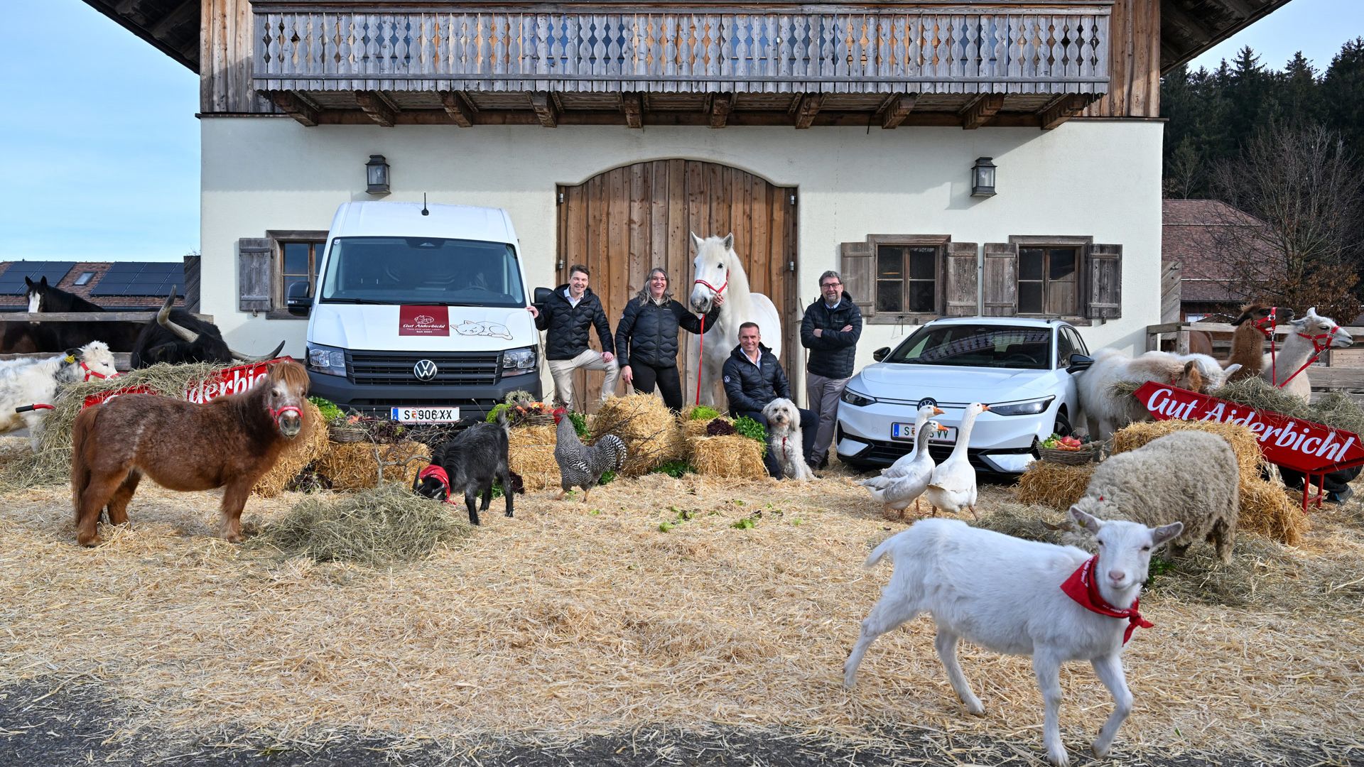 Mehrere Menschen und zahlreiche Tiere, darunter Ziegen, Hühner und Schafe, stehen vor einem alpinen Bauernhaus mit Holzbalkon. Im Hintergrund sind zwei Fahrzeuge geparkt, ein weißer Lieferwagen und ein helles Auto. Einige der Tiere tragen rote Schärpen mit der Aufschrift "Gut Aiderbichl".